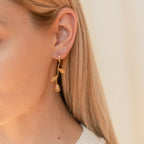 Close-up of a woman's ear adorned with Dangling Rose Huggies—gold, Venetian-style earrings with a rose design—elegantly complementing her long blonde hair.