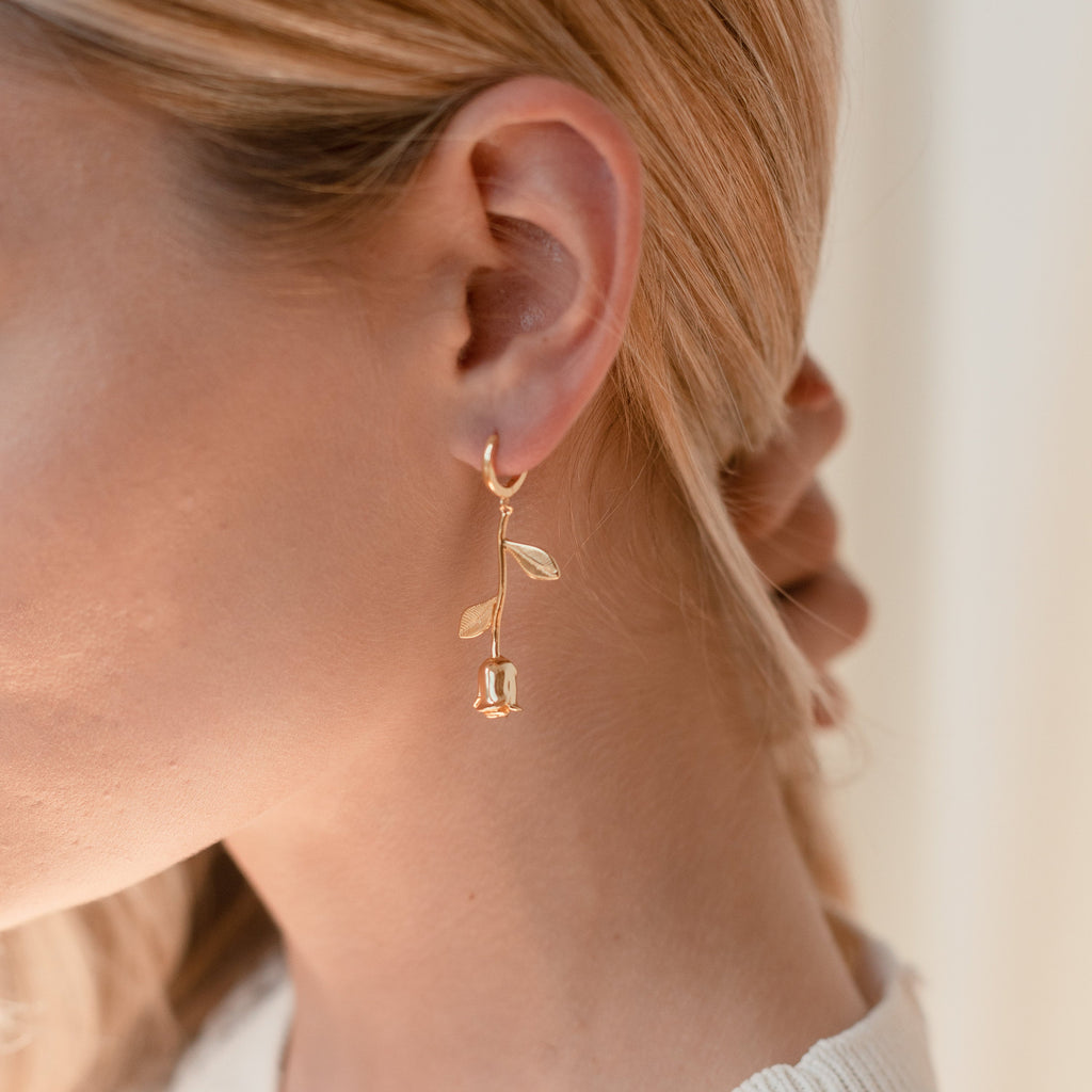 Close-up of a woman wearing Dangling Rose Huggies—a romantic, gold flower-shaped earring—against a light background.