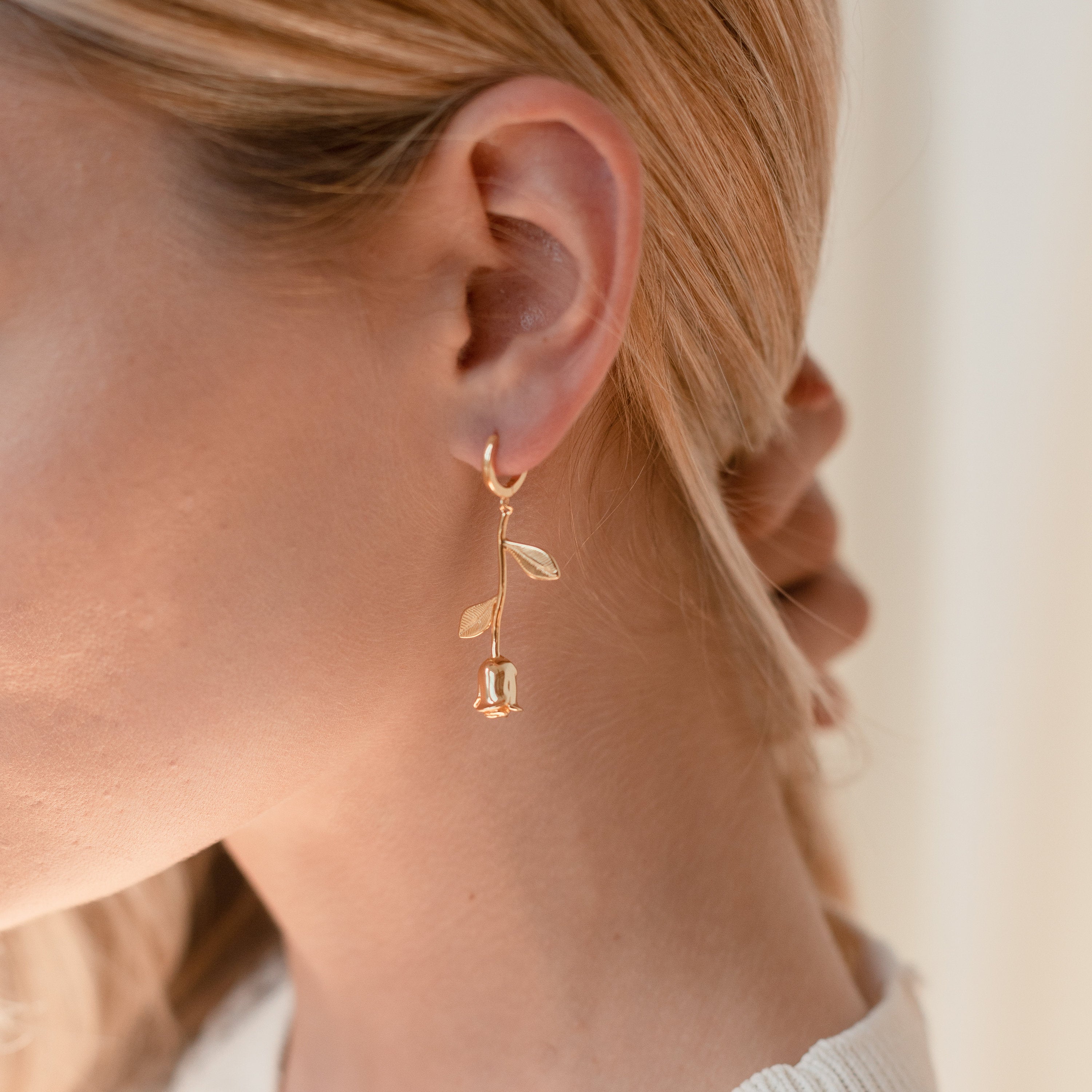 Close-up of a woman wearing Dangling Rose Huggies—a romantic, gold flower-shaped earring—against a light background.