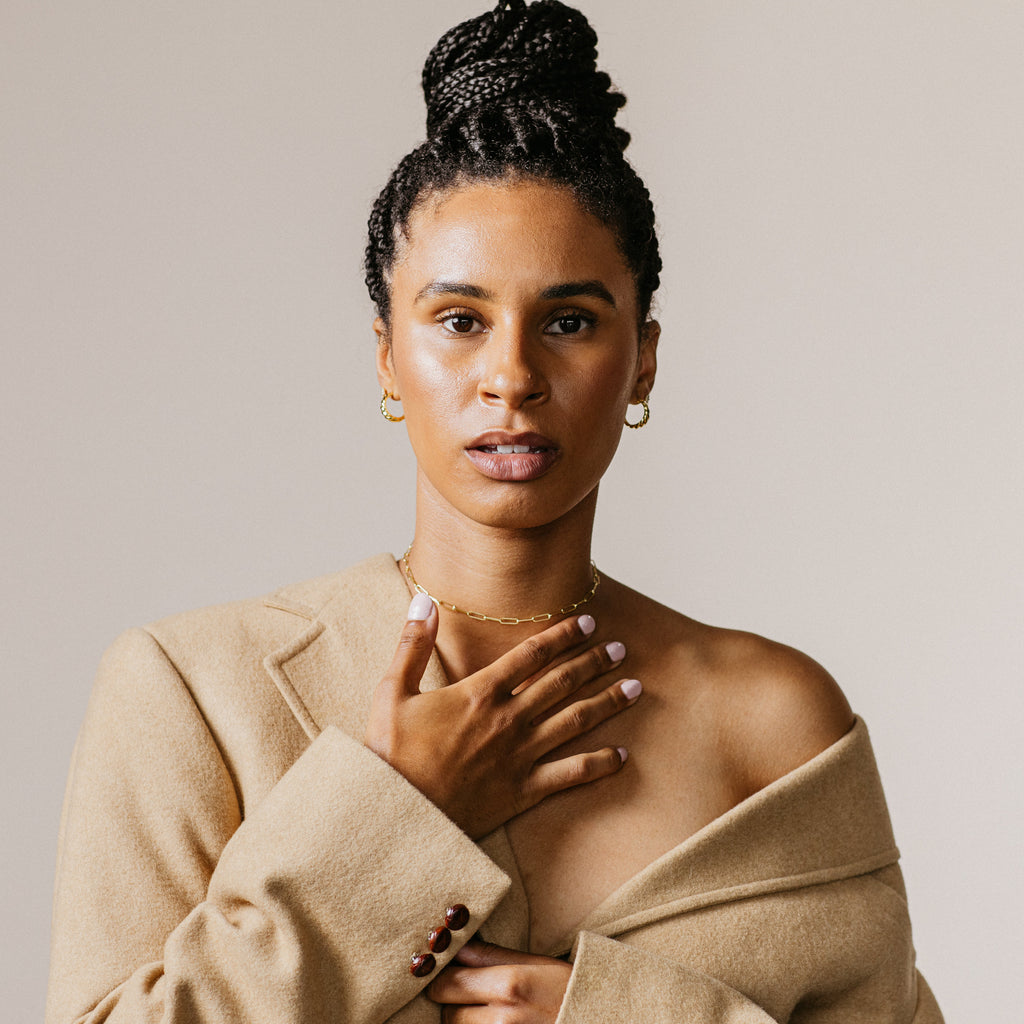 Woman with braided hair in a tan coat, hand on chest, wears Croissant Hoops and looks confidently at the camera against a plain background.