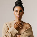 Woman with braided hair in a tan coat, hand on chest, wears Croissant Hoops and looks confidently at the camera against a plain background.