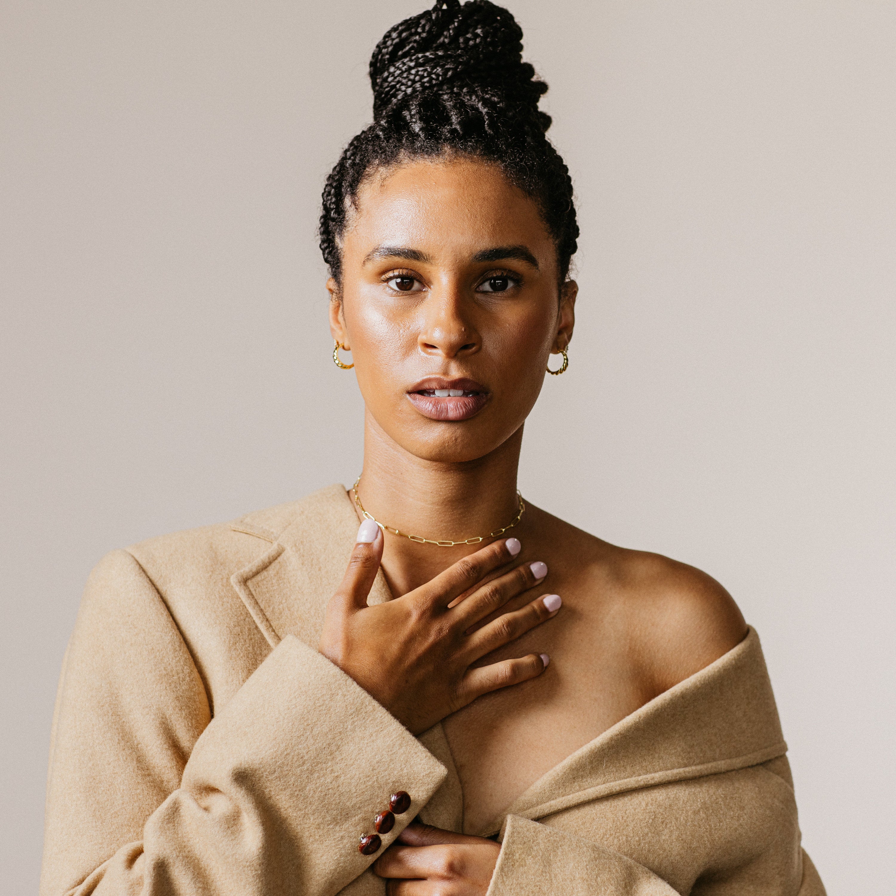 Woman with braided hair in a tan coat, hand on chest, wears Croissant Hoops and looks confidently at the camera against a plain background.