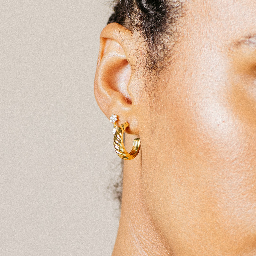 Close-up of a person’s ear wearing Croissant Hoops, small gold hoops with twist detailing, alongside a sparkling diamond stud.
