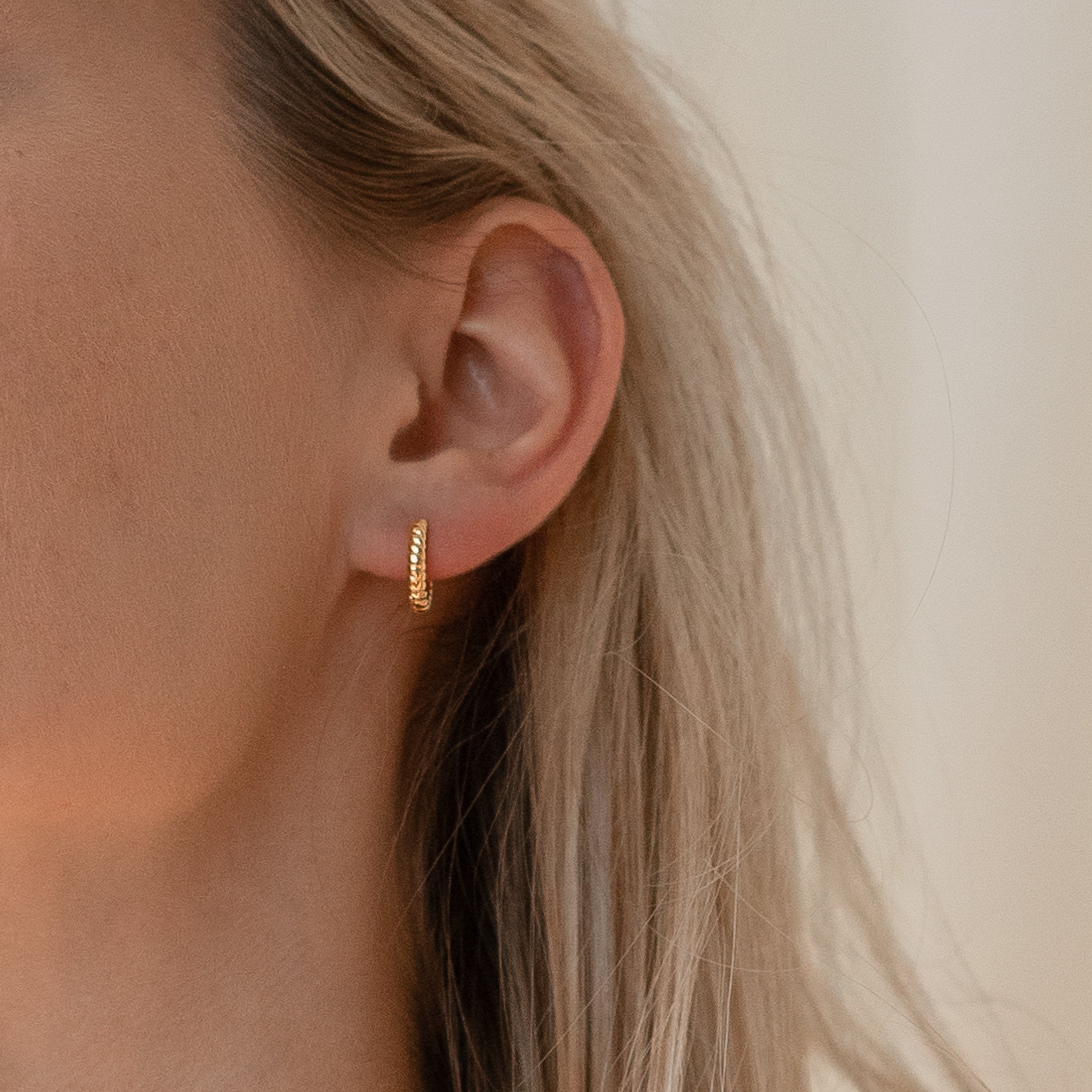A close-up of a woman's ear adorned with Thea Twisted Huggies in 18K Gold.
