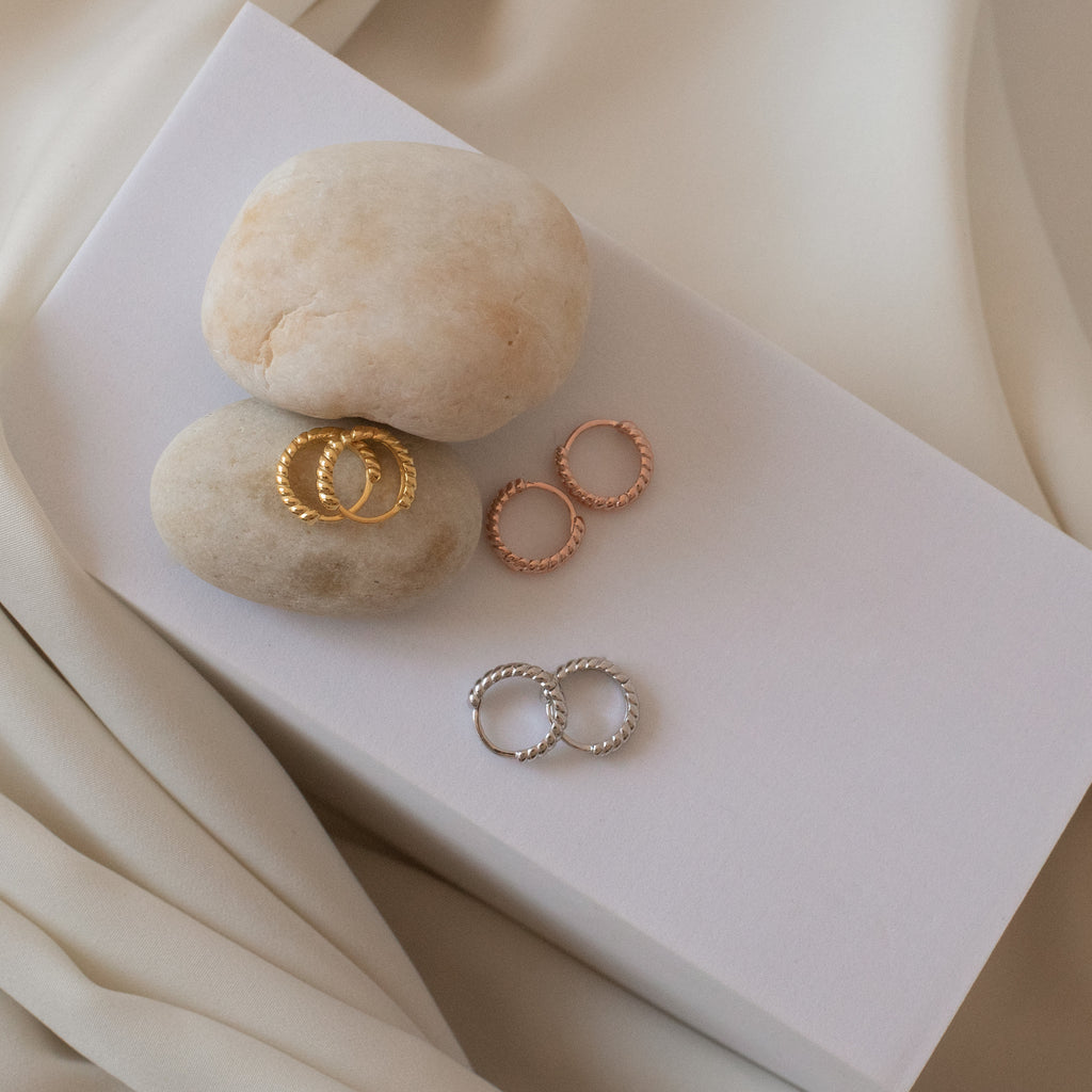 The Thea Twisted Huggies in Rose Gold—a minimalist, textured hoop earring—shown on stones and white fabric, perfect for everyday elegance.