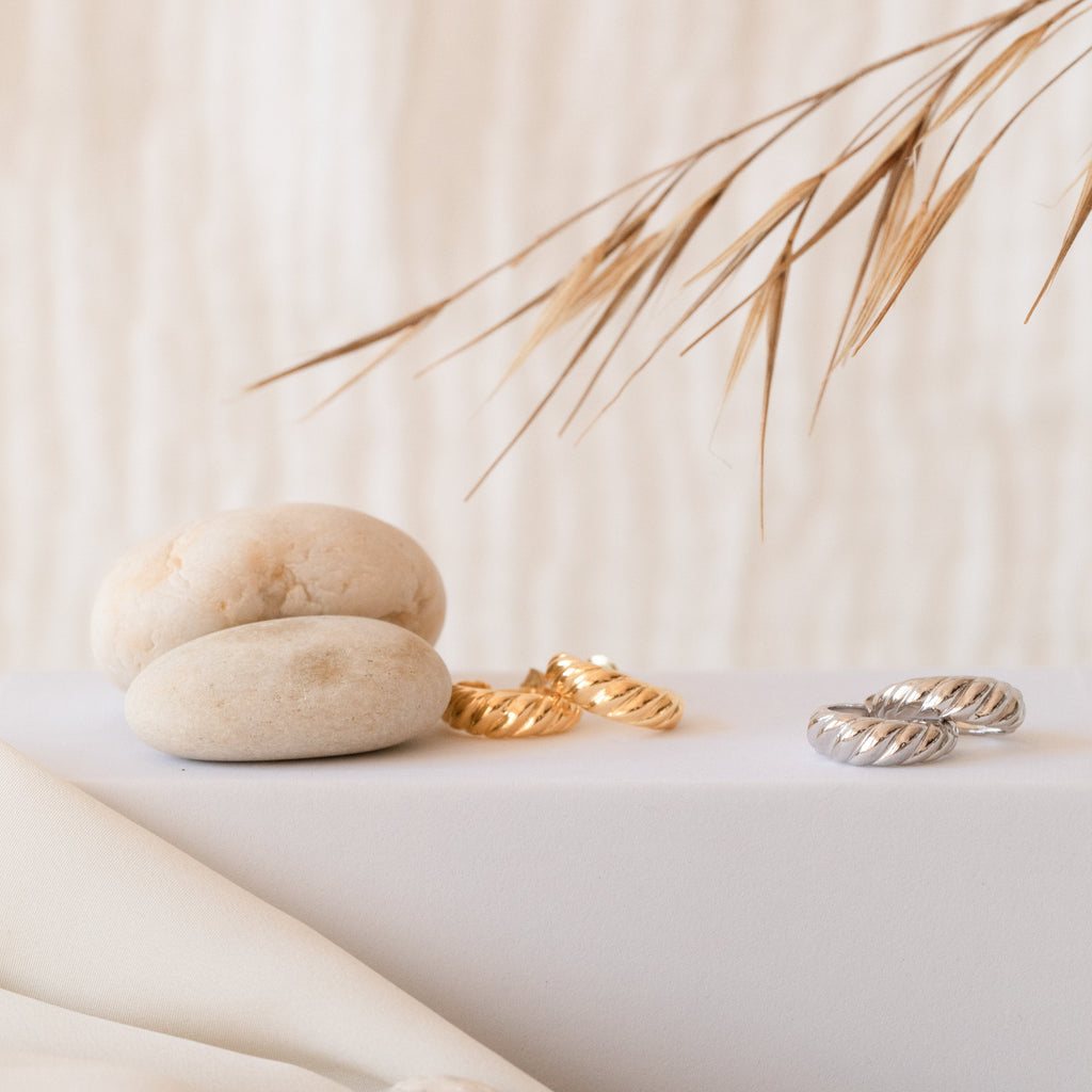 Croissant Hoops with textured, twisted design rest beside smooth stones and dried grass on a light, minimalist background.