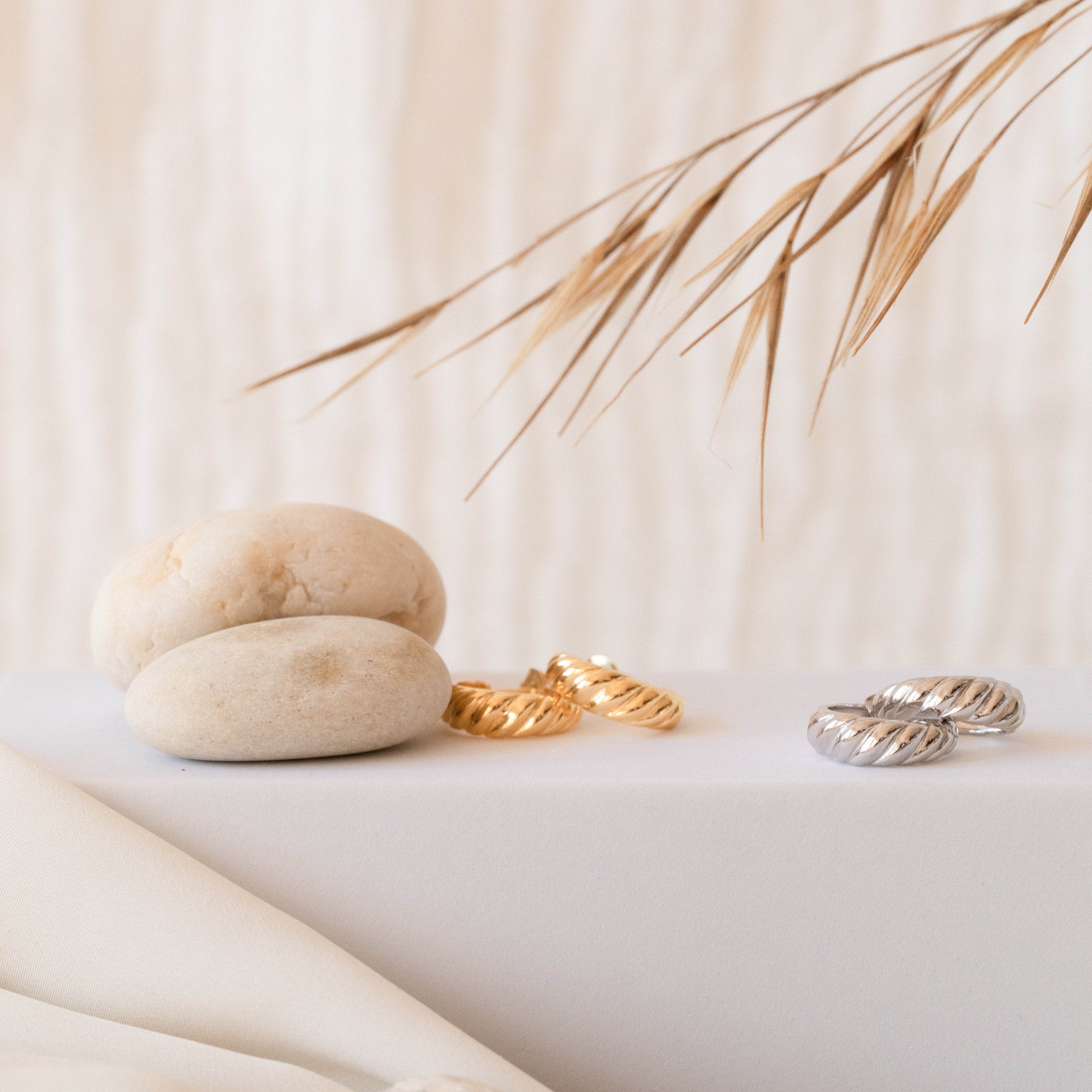 Croissant Hoops with textured, twisted design rest beside smooth stones and dried grass on a light, minimalist background.