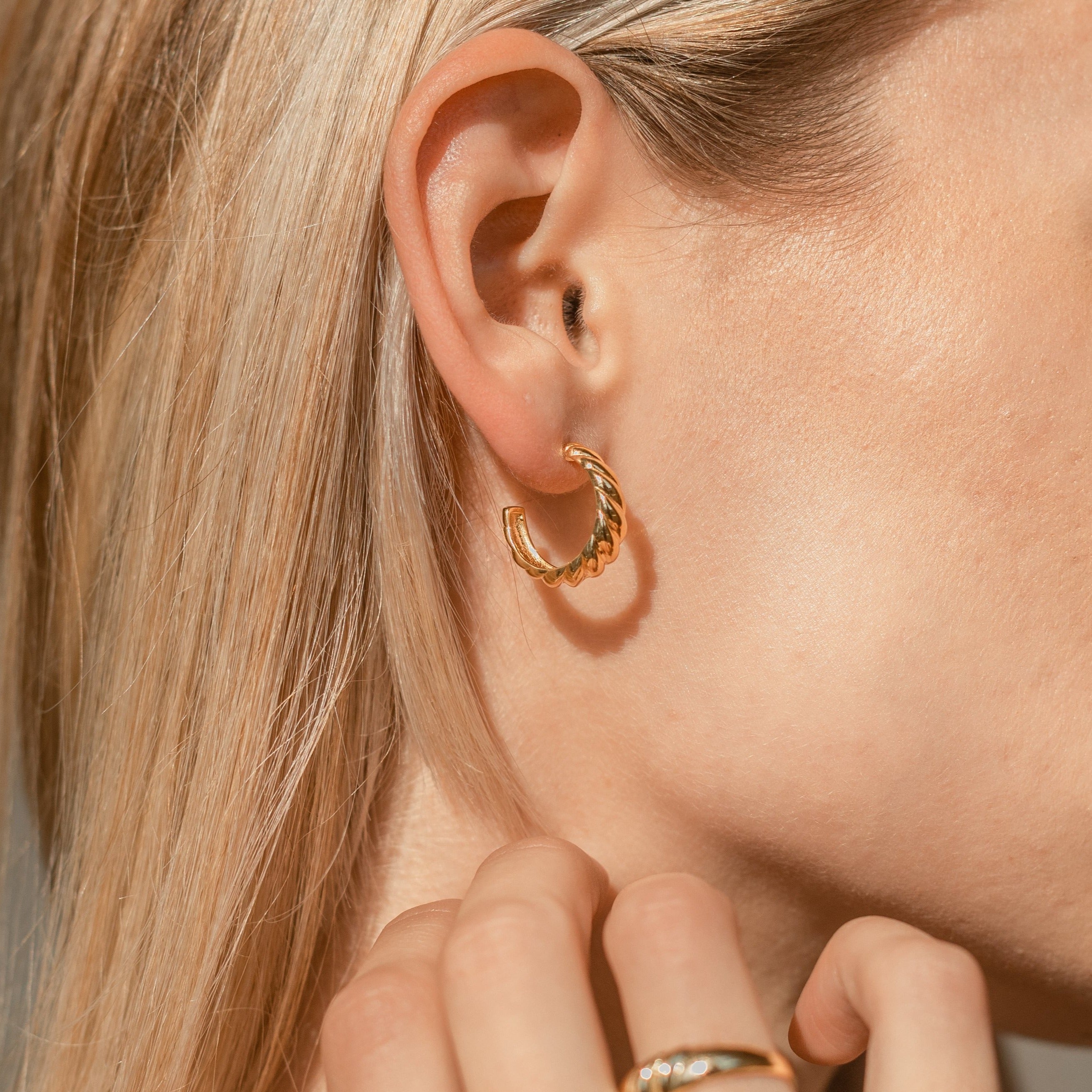 Close-up of a woman’s ear wearing gold Croissant Hoops with twist detailing, her blonde hair cascading and hand near her face.