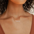 Close up of a woman wearing a gold necklace with the word “mama” spelled out in sparkling pavé script letters. The pendant hangs from a fine chain and rests just above the collarbone, styled with a rust colored tank top.
