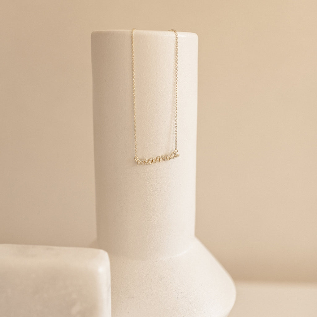 The Pave Mama Script Necklace features a sparkling cursive pendant, elegantly displayed on a smooth, cream-colored ceramic vase—an ideal piece of motherhood jewelry and the perfect gift for Mom.