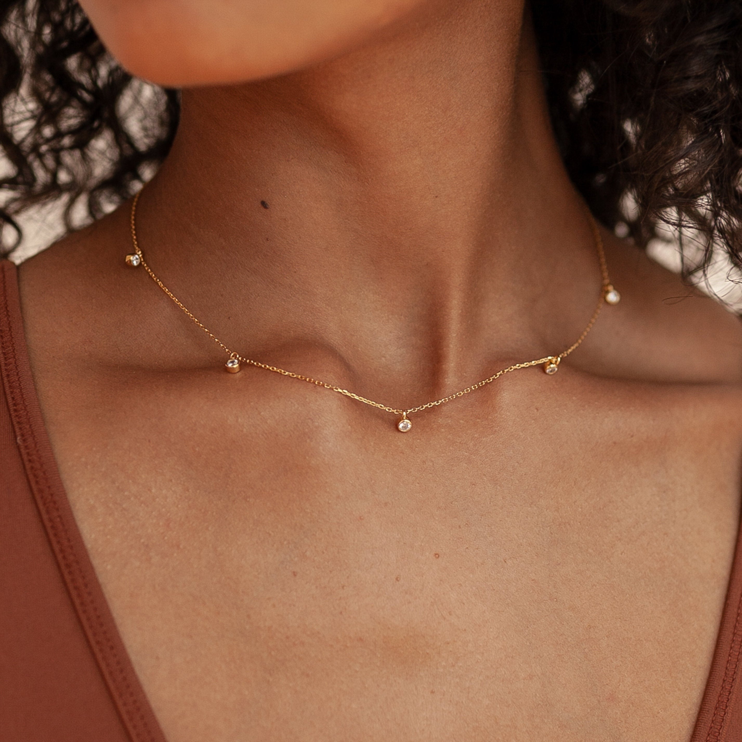 Minimalist gold necklace with five bezel set crystal charms spaced along a fine chain, worn above the collarbone with a v neck top.