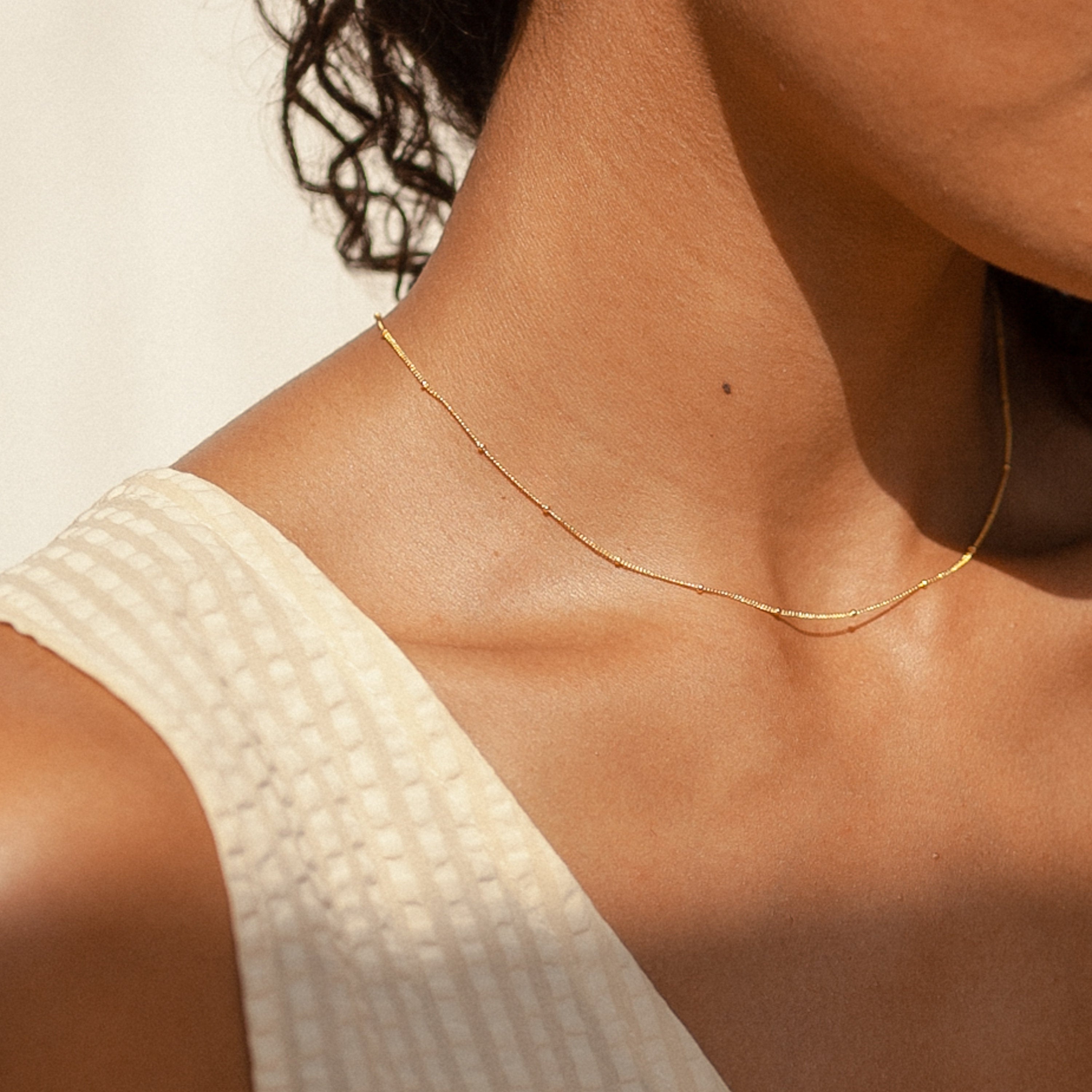 The gold Satellite Necklace worn close to the neck by a model in a cream textured top, under warm natural light.