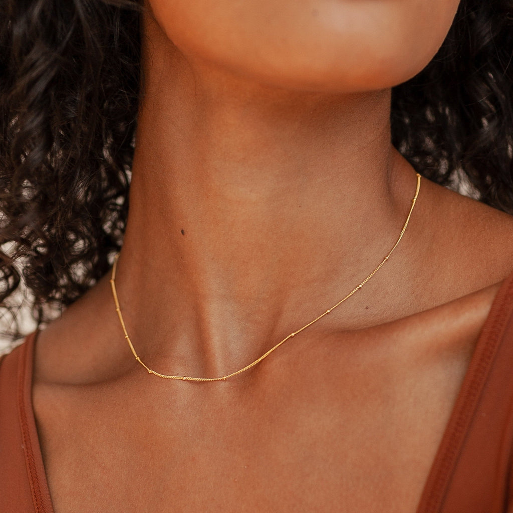 A gold Satellite Necklace worn by a model in a brown top, the chain forming a subtle wave across the collarbone.