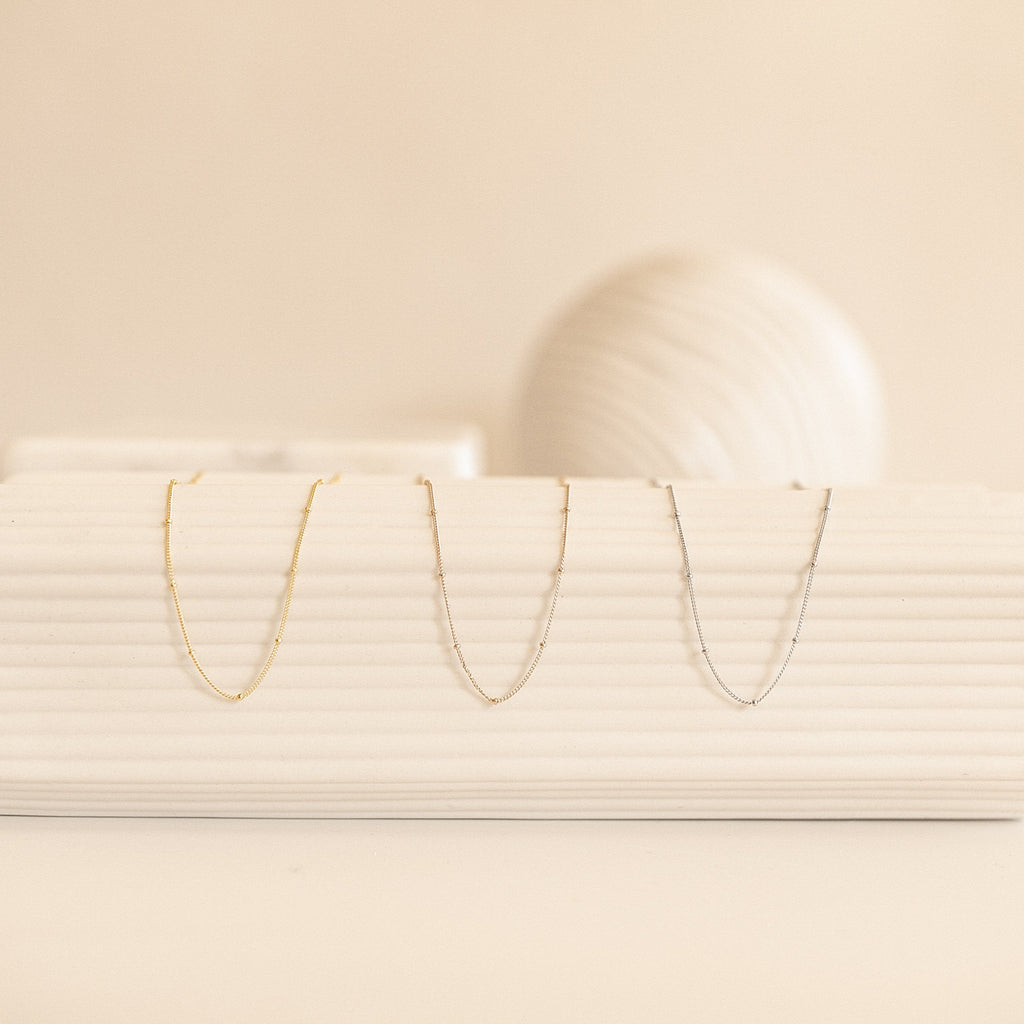 Three Satellite Necklaces—yellow gold, rose gold, and white gold—are displayed hanging neatly from a white ribbed ceramic display.