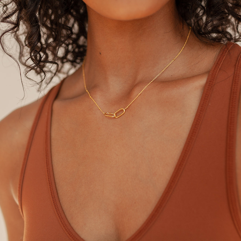 A woman wears the Linked Pendant Necklace, a delicate geometric gold chain, paired with a rust-colored tank top.