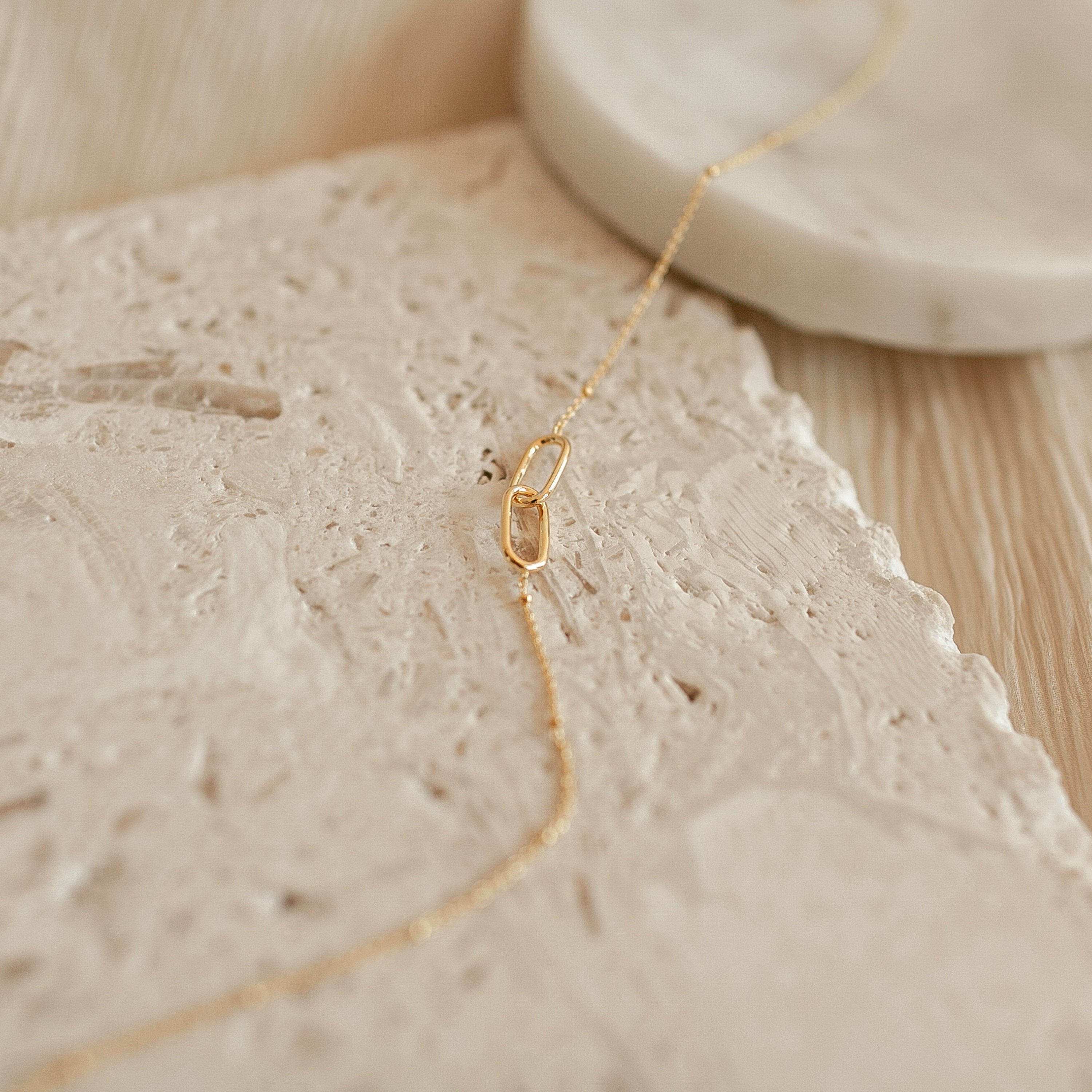 The Linked Pendant Necklace in Rose Gold features a delicate chain with an oval-linked pendant, elegantly displayed on a textured beige stone surface.