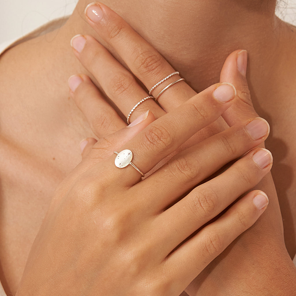 Close-up of manicured hands wearing minimalist silver rings and the Constellation Oval Ring, fingers crossed gracefully at the neck.