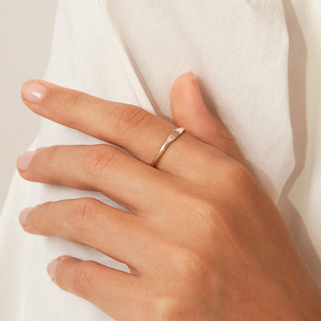 A hand with neatly manicured nails wears the Skinny Zodiac Ring, a simple silver stacking band, against a white fabric background.