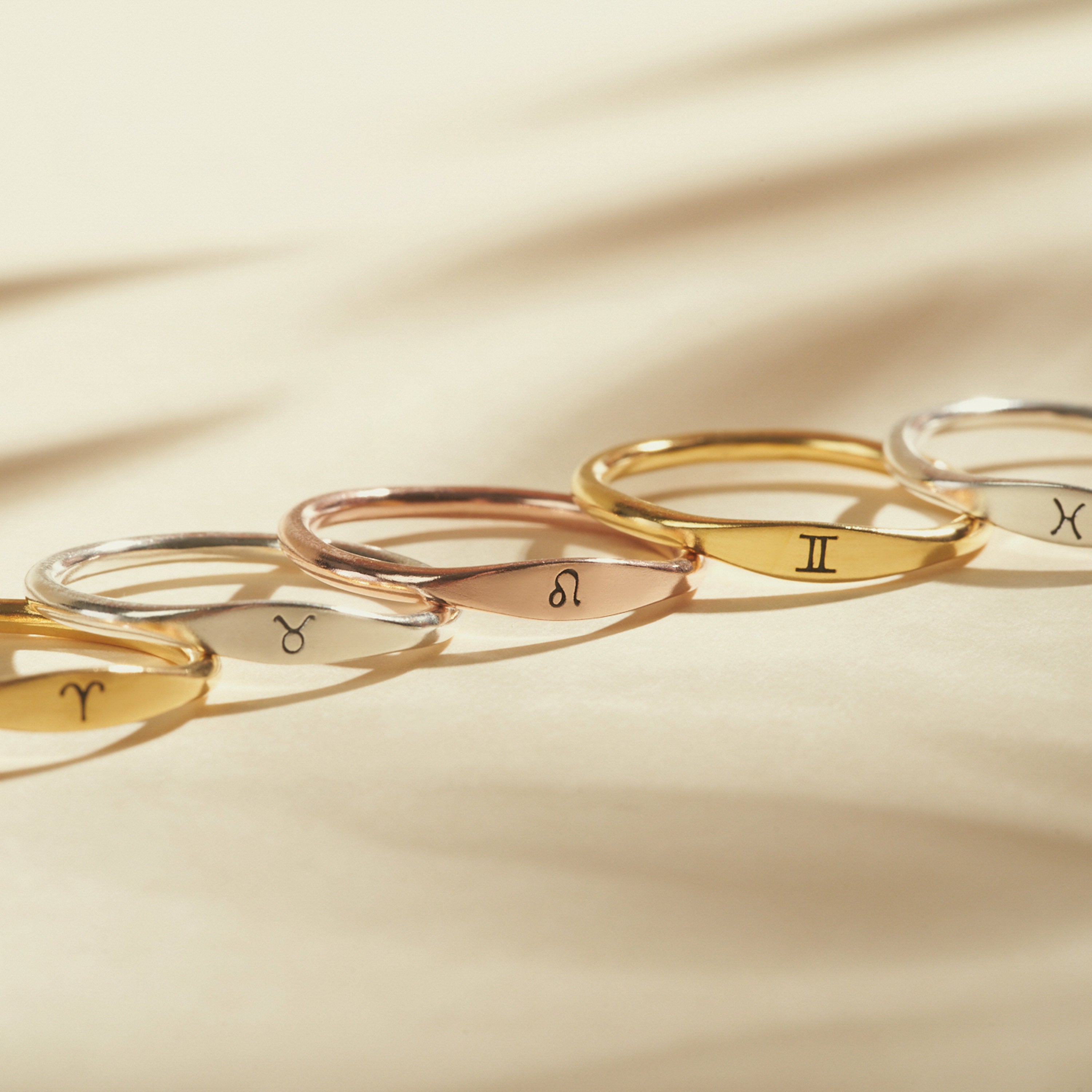 Five Skinny Zodiac Rings with engraved zodiac symbols are arranged in a row on a beige surface, illuminated by soft lighting.