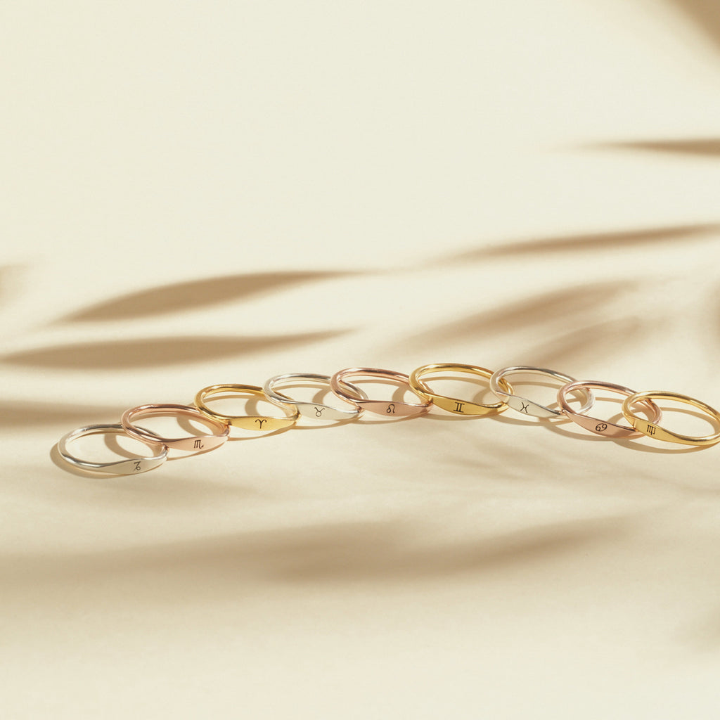 Nine Skinny Zodiac Rings in gold and silver are neatly arranged on a beige surface, with soft shadows in the background.