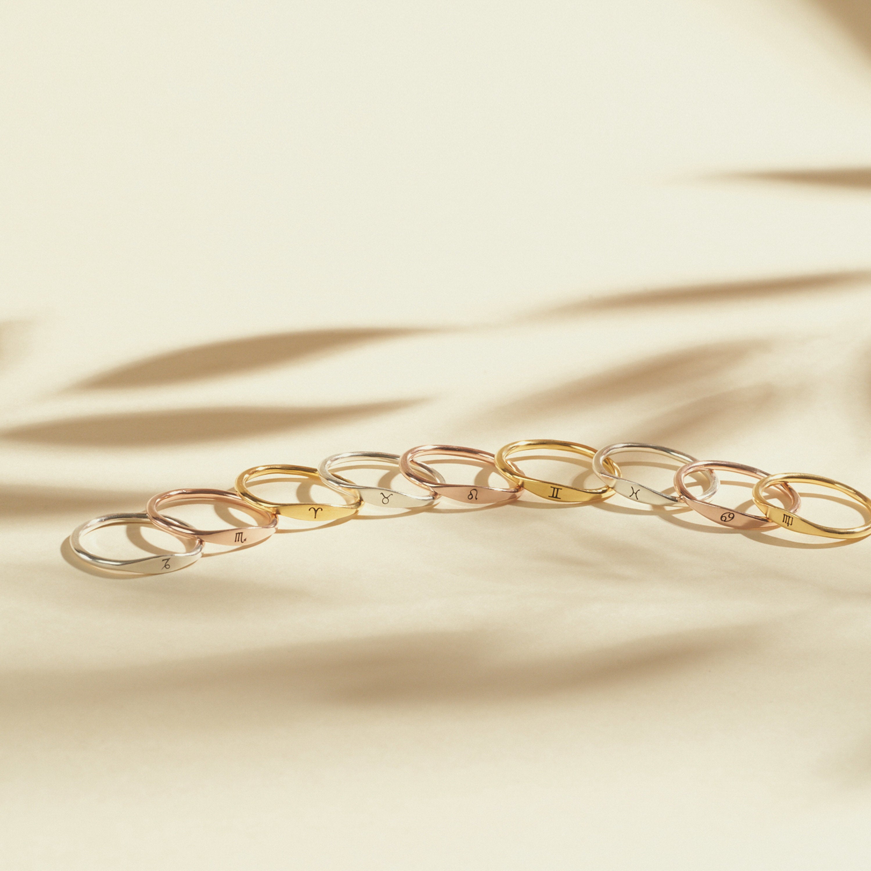 Nine Skinny Zodiac Rings in gold and silver are neatly arranged on a beige surface, with soft shadows in the background.