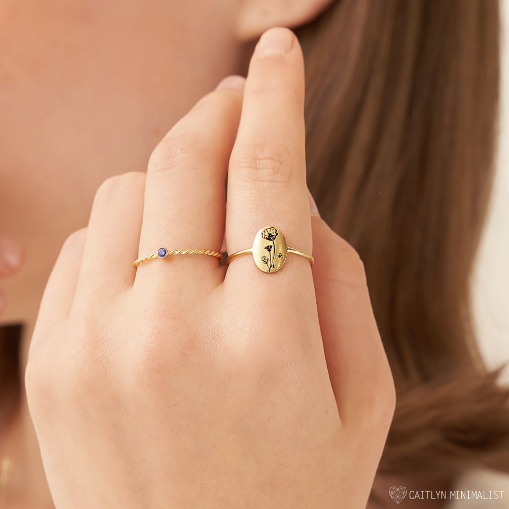A hand wearing two gold rings—one with a small stone and the other the Birth Flower Oval Ring, showcasing an engraved floral design for a personalized jewelry touch.