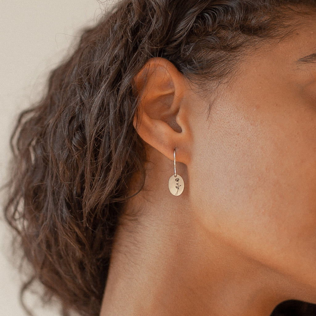 Close-up of a woman’s ear wearing Dangling Birth Flower Earrings—silver oval statement pieces with a small leaf design, perfect for those who love subtle yet elegant jewelry.
