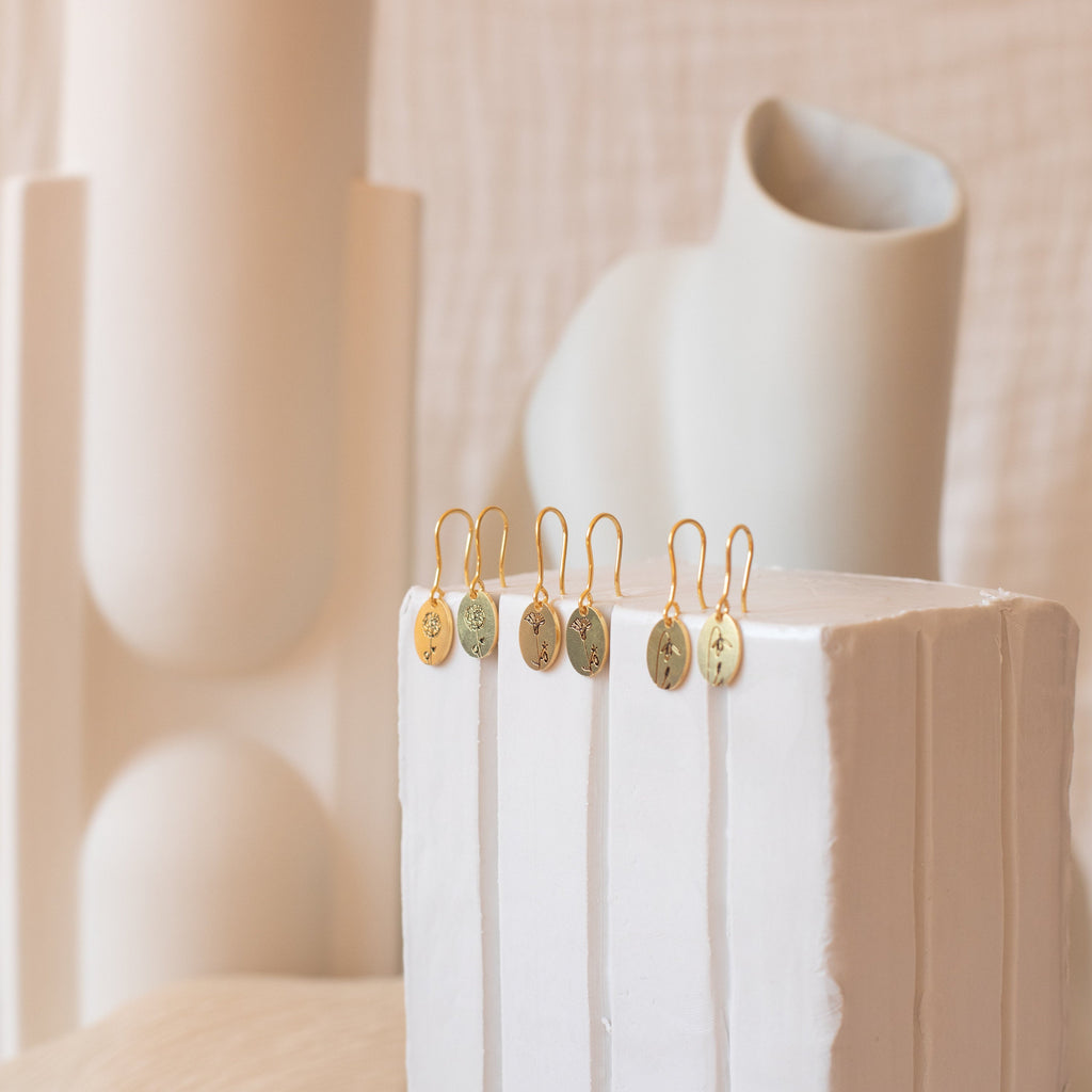 Five Dangling Birth Flower Earrings are displayed on a white textured block with abstract cream-colored sculptures behind them. Add a personalized, elegant touch to any outfit with these stunning earrings.