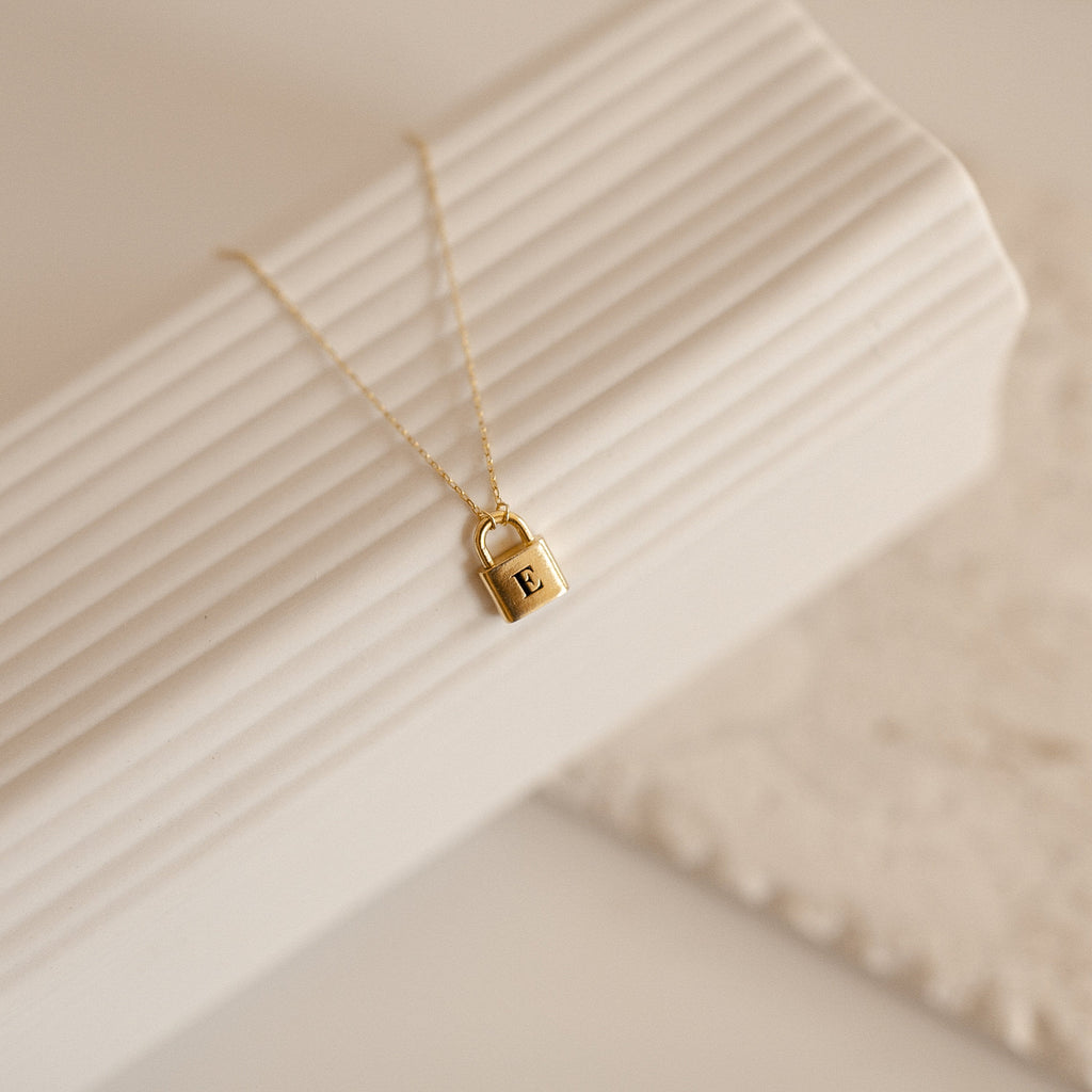 Padlock Necklace: Gold necklace featuring a padlock pendant engraved with the letter E, perfect for adding a chic touch to any look. Shown on a textured white background.