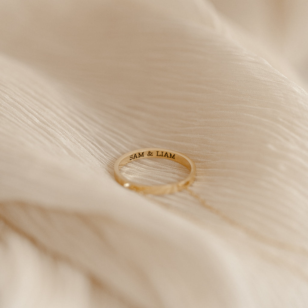 The Inner Circle Necklace, engraved with SAM & LIAM, rests on a soft, cream-colored fabric background, evoking the sentiment of a treasured amulet.