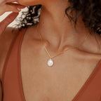 A woman in a rust-colored top wears the Sadie Pearl Necklace—a gold baroque chain featuring a teardrop-shaped freshwater pearl pendant—making it an elegant bridesmaid gift.