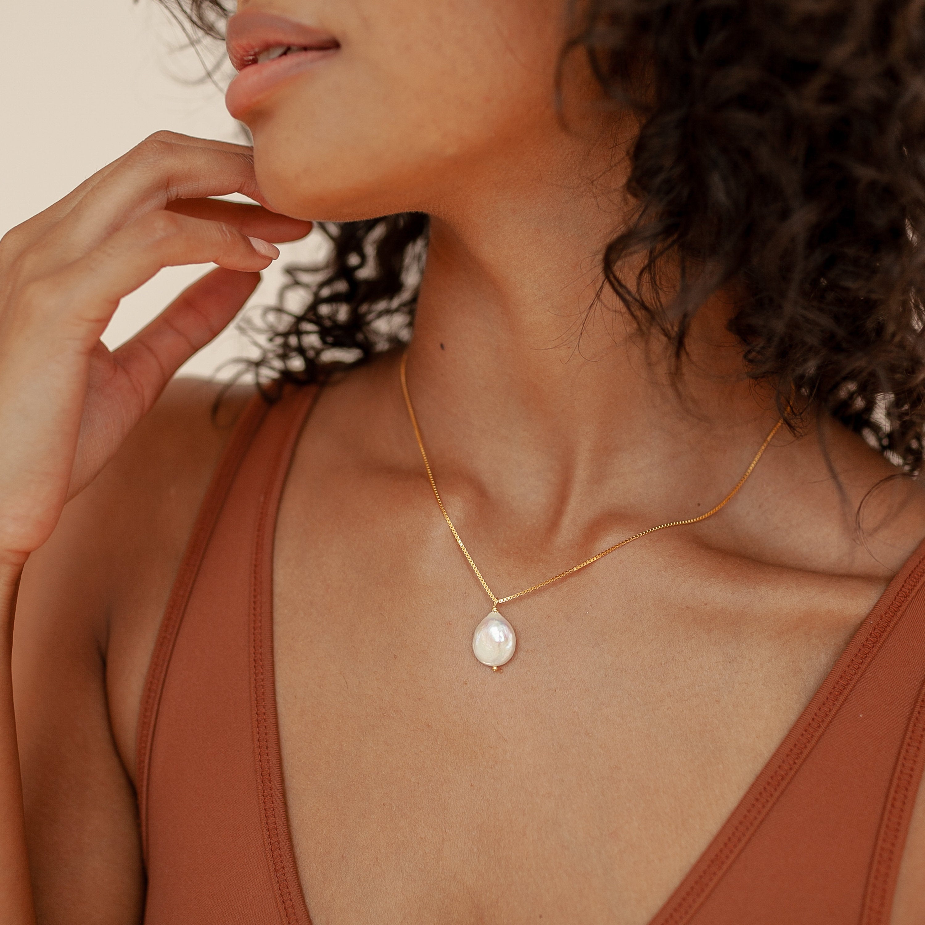 A woman in a brown top gently touches her face while wearing the Sadie Pearl Necklace—a gold baroque chain with a pearl pendant—creating an elegant look ideal for bridesmaids gifts.