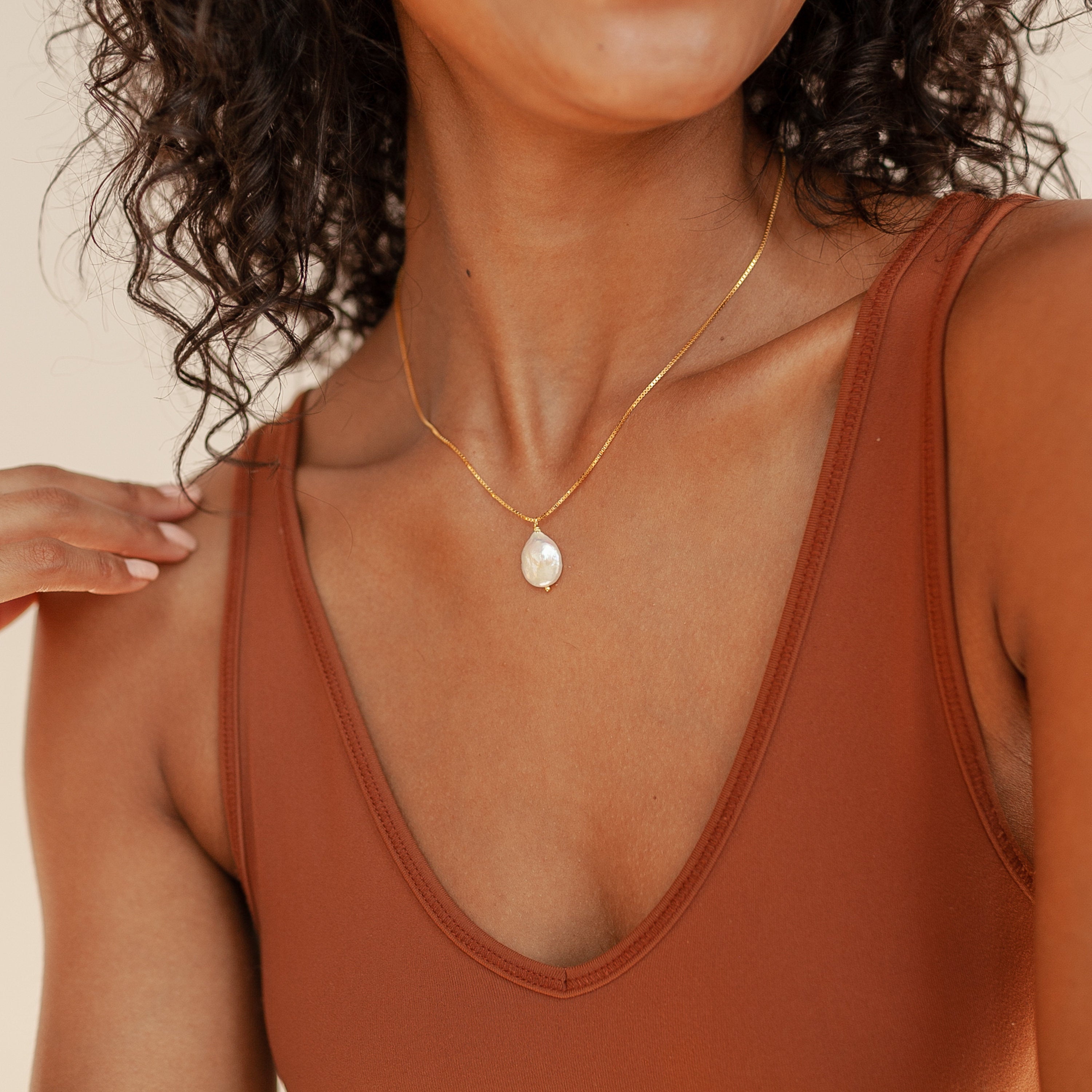 Woman wearing a brown tank top and the Sadie Pearl Necklace, a gold baroque necklace with a round pendant, hand touching her shoulder.