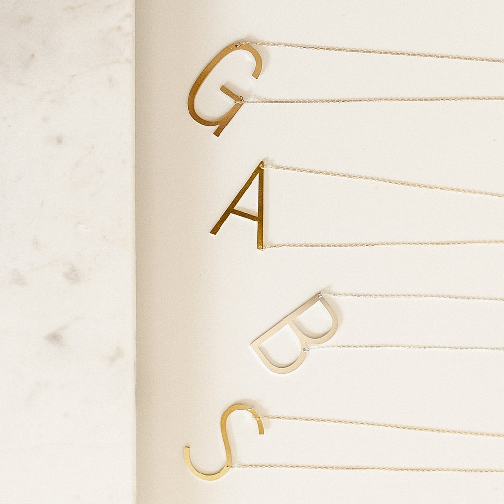 Four Large Sideways Initial Necklaces in Rose Gold—featuring bold G, A, B, and S letter charms—are arranged on a white surface for a striking personalized statement.