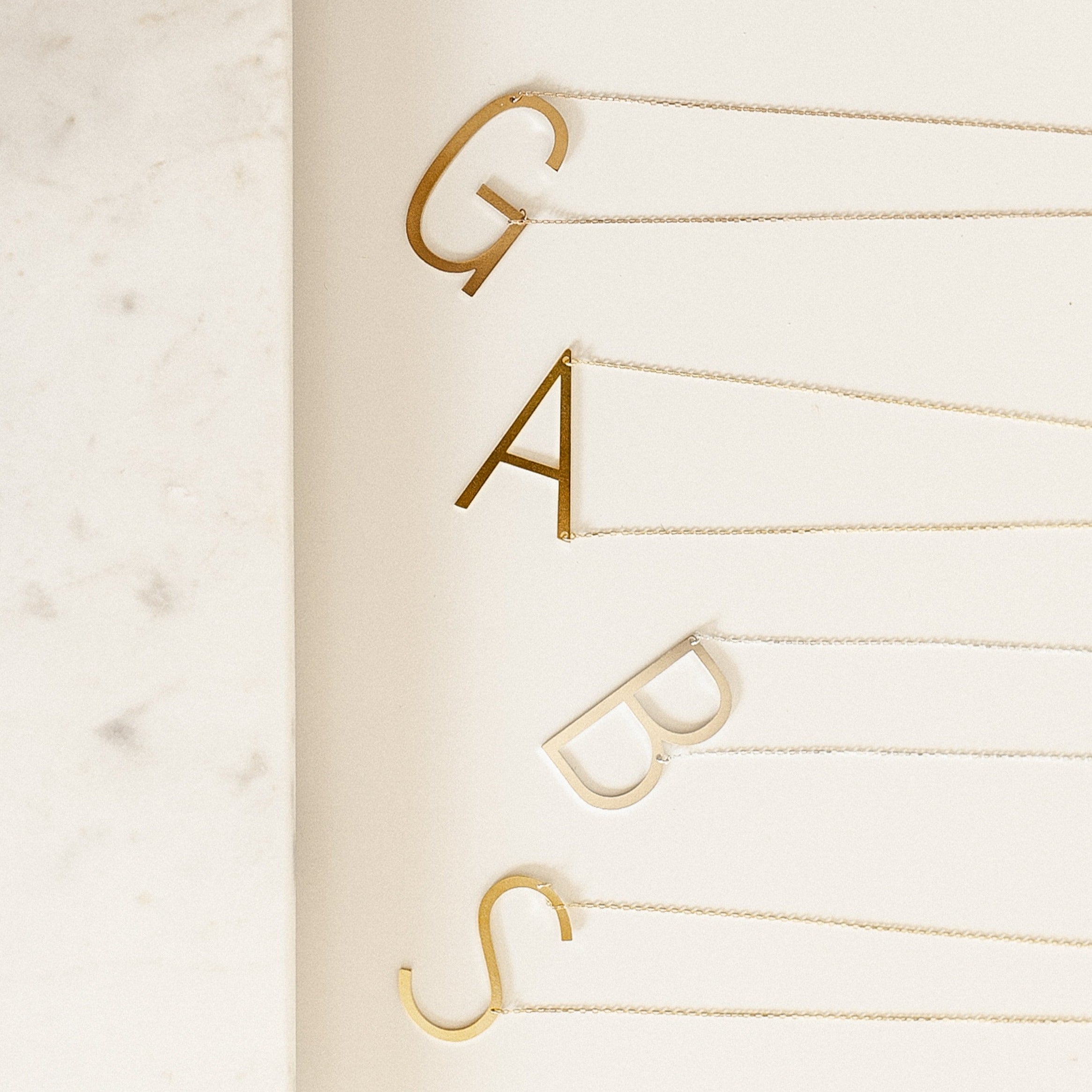 Four Large Sideways Initial Necklaces in Rose Gold—featuring bold G, A, B, and S letter charms—are arranged on a white surface for a striking personalized statement.