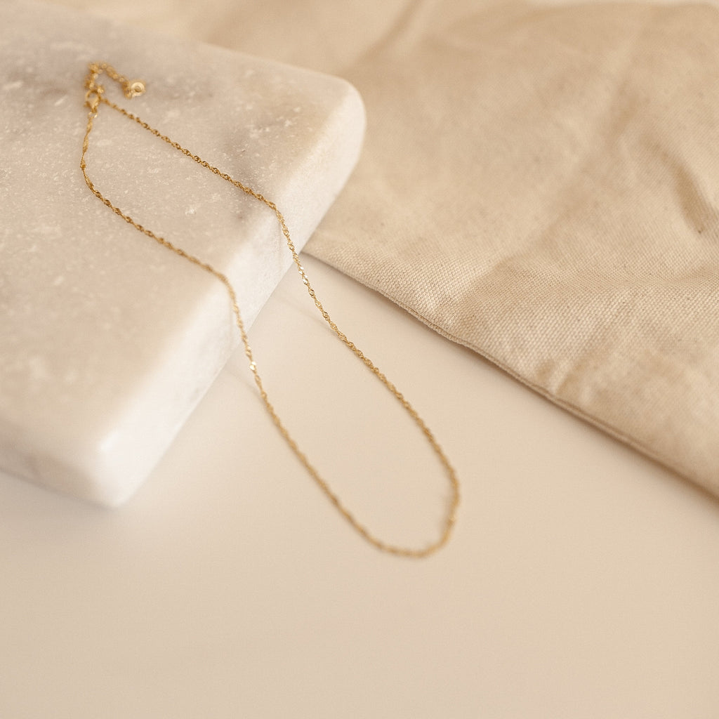 The Singapore Twist chain displayed in a flat lay over marble and linen.