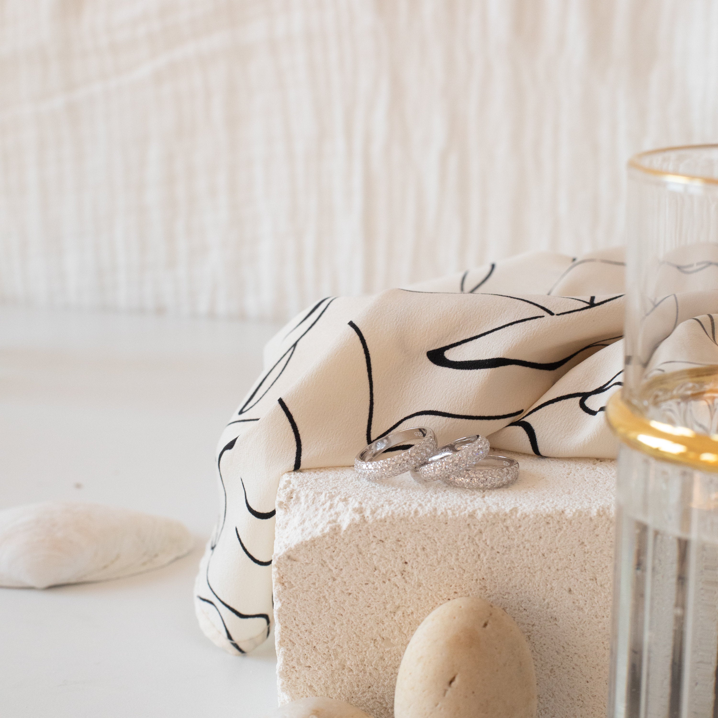 The Pave Dome Ring, along with two other silver rings, rests on a textured block accented by patterned cloth, glass, and stones in a minimal setting.