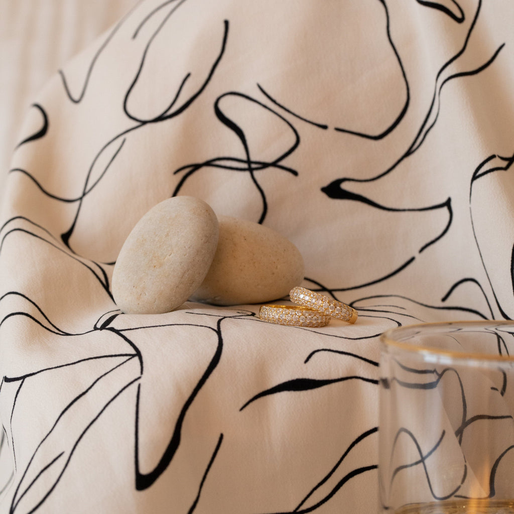 A Pave Dome Ring, a signature timepiece, two smooth stones, gold rings—including a dainty band—and a glass are displayed on a patterned white cloth with black abstract lines.