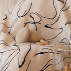 A Pave Dome Ring, a signature timepiece, two smooth stones, gold rings—including a dainty band—and a glass are displayed on a patterned white cloth with black abstract lines.