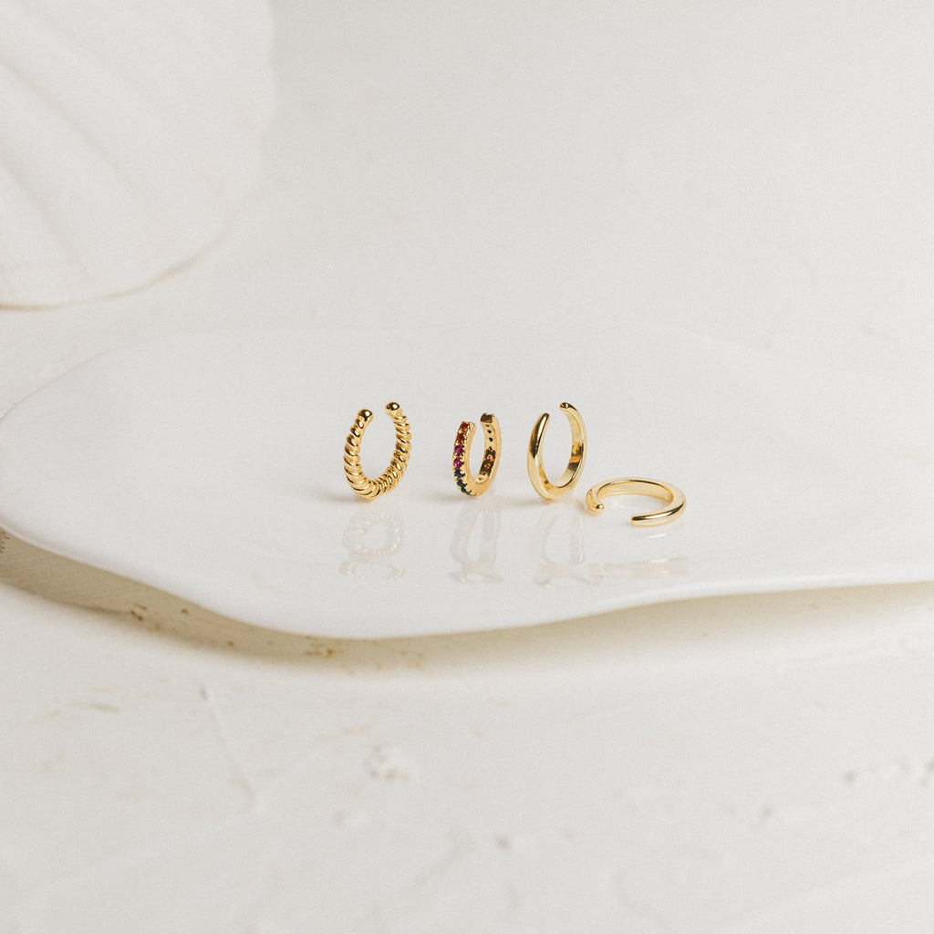 The Cleo Cuffs set features four small gold hoops, including stylish ear cuffs and cartilage earrings, displayed on a white ceramic dish against a neutral background.