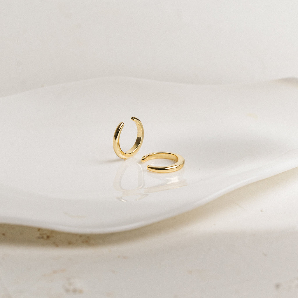 Two Cleo Cuffs, small gold hoop ear cuffs, rest on a wavy white ceramic dish against a neutral background, providing a stylish option for non-pierced earrings.