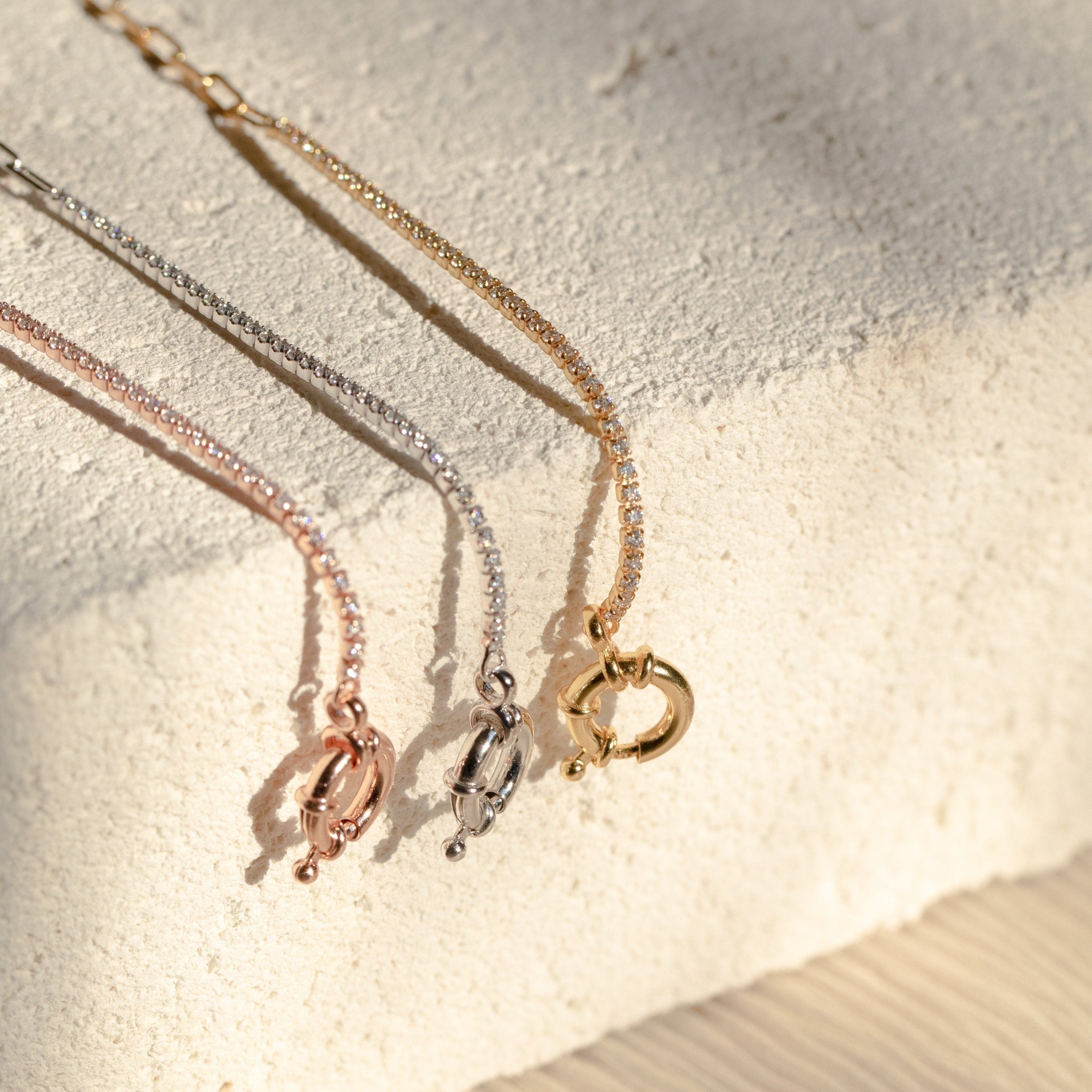 The Klara Tennis Bracelet—a sleek, minimalist piece in rose gold, silver, or gold—makes a perfect addition to any jewelry collection and is displayed on a textured, light-colored surface.