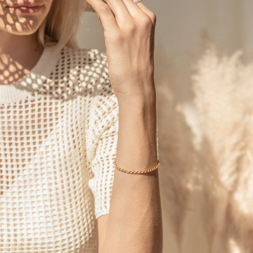 A woman in a white textured top wears the Rope Chain Bracelet, featuring a twisted gold design, as sunlight casts patterns on her skin.