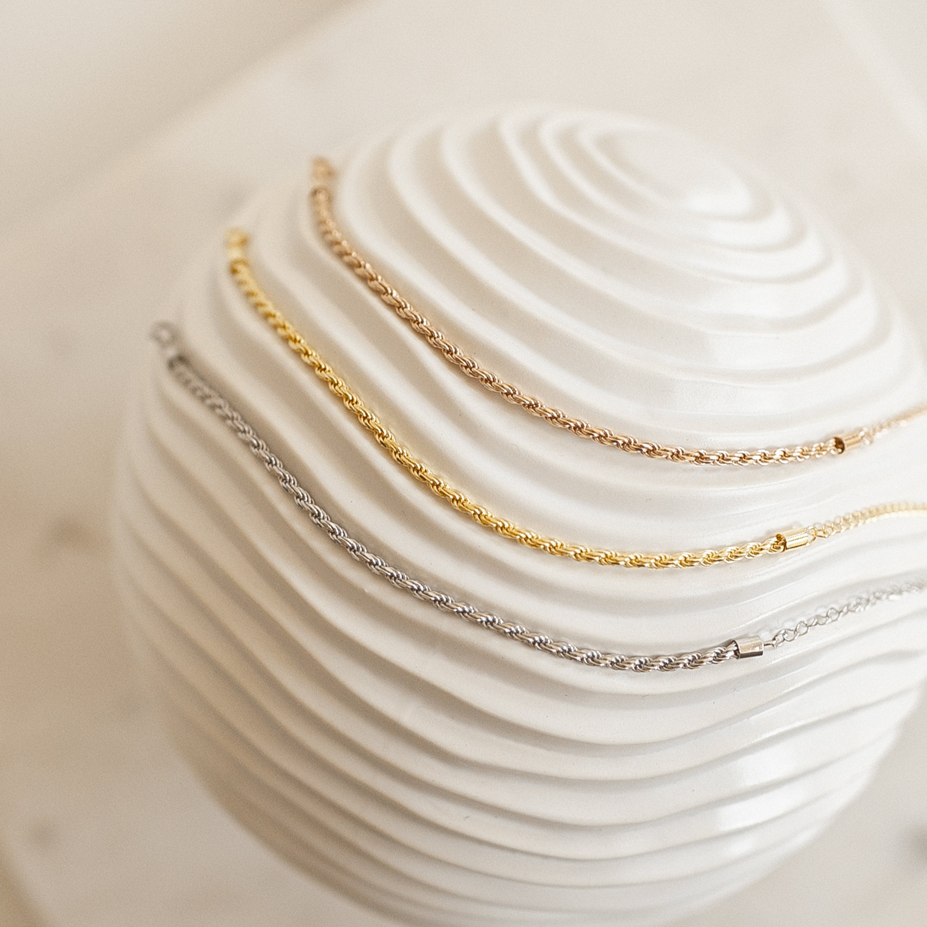Three delicate chain bracelets—one twisted, a Rope Chain Bracelet, and one rose gold—are displayed on a white textured sphere.