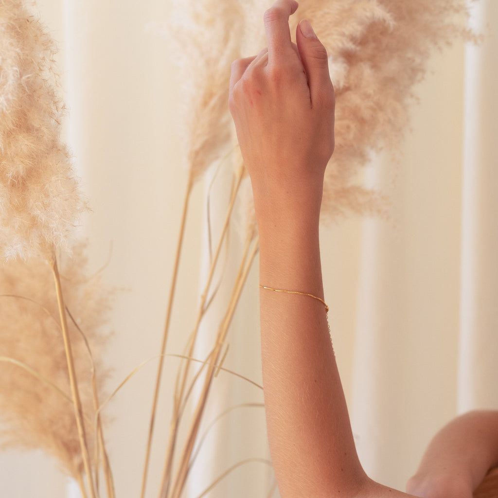 A raised hand wears the Box Chain Bracelet with a polished finish and slender gold links, framed by soft beige pampas grass.
