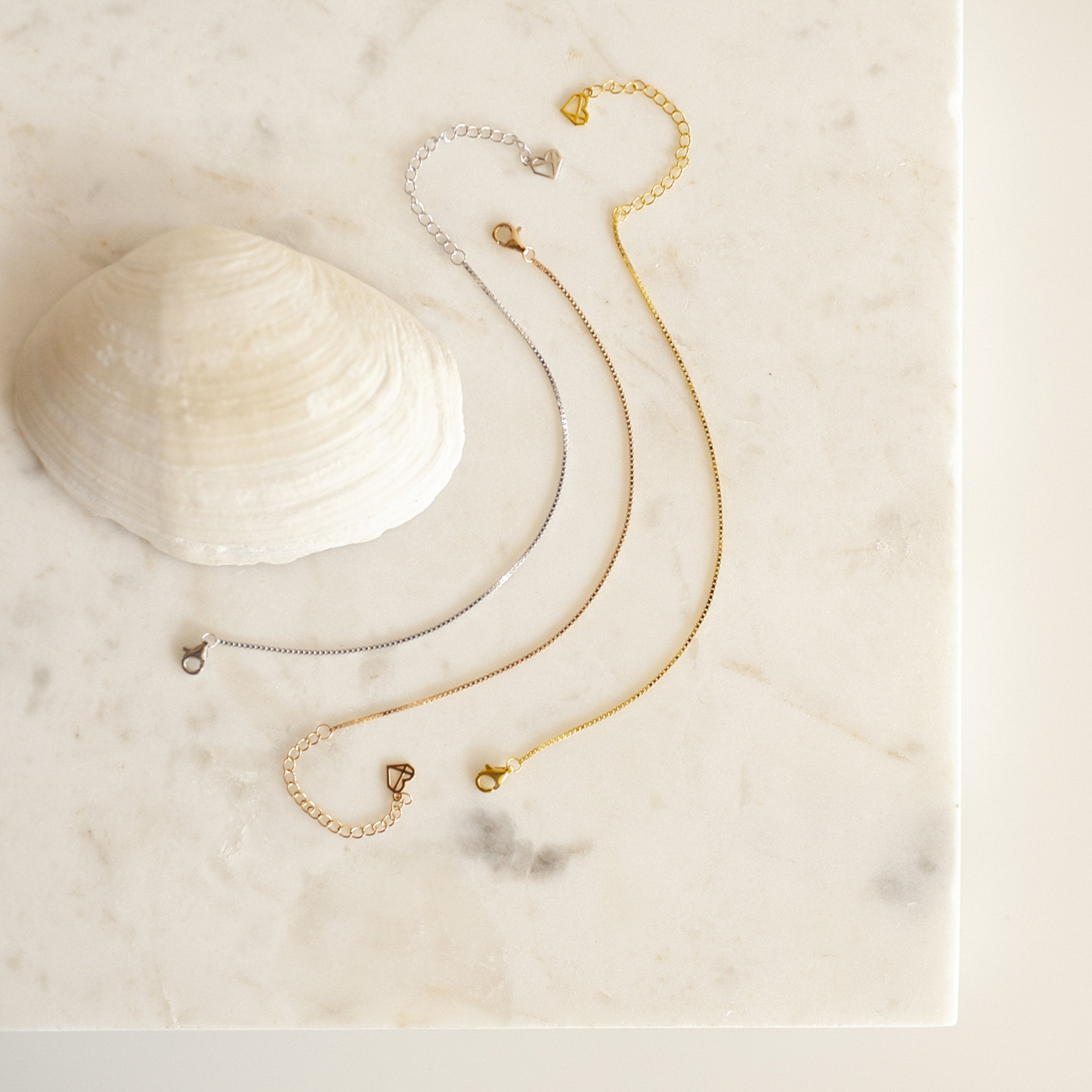 The polished Box Chain Bracelet in gold and silver rests beside a seashell on a marble surface, paired with two delicate chain bracelets.