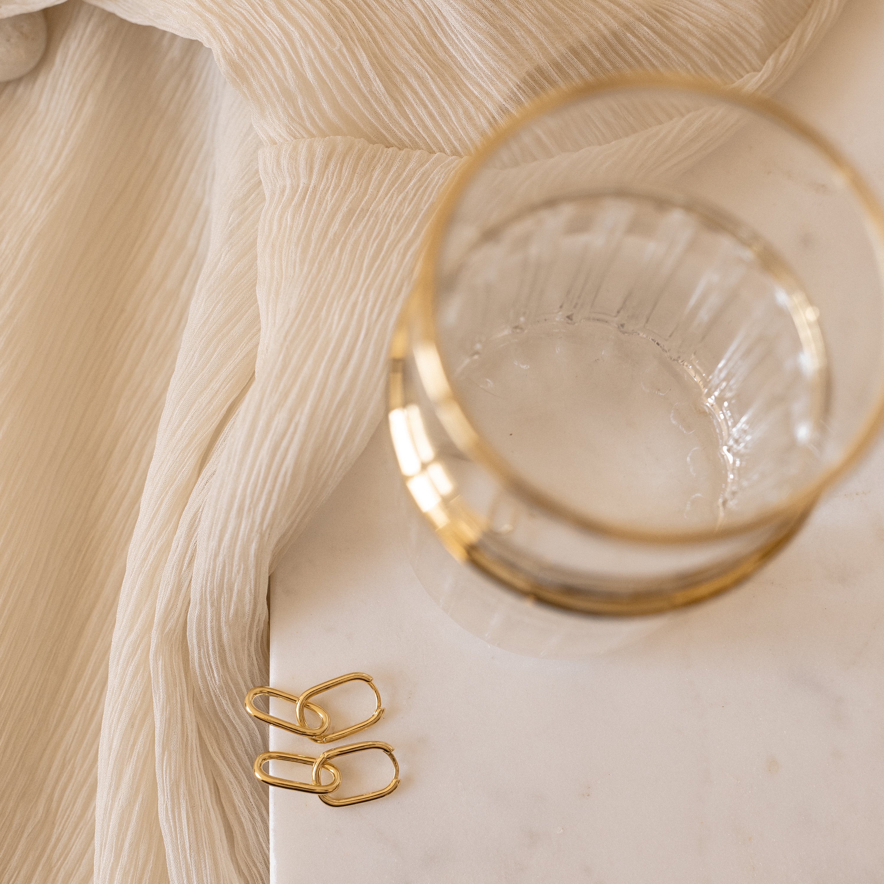 The Amanda Link Earrings in 18K Gold are displayed on a white marble surface with a cream-colored fabric background.