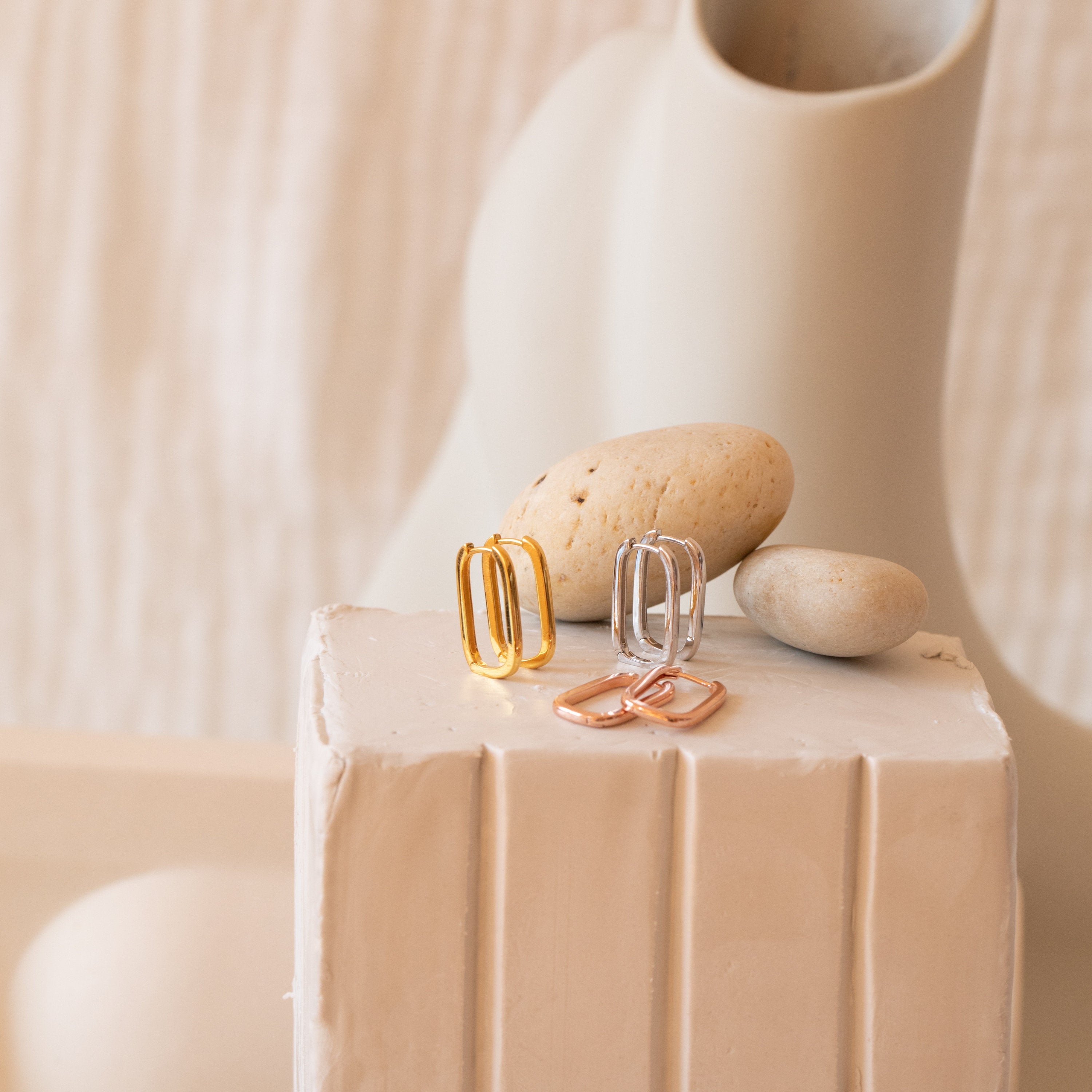 The Nina Link Earrings in Rose Gold are displayed on a white textured pedestal, offering a modern twist on classic rectangular hoop earrings.