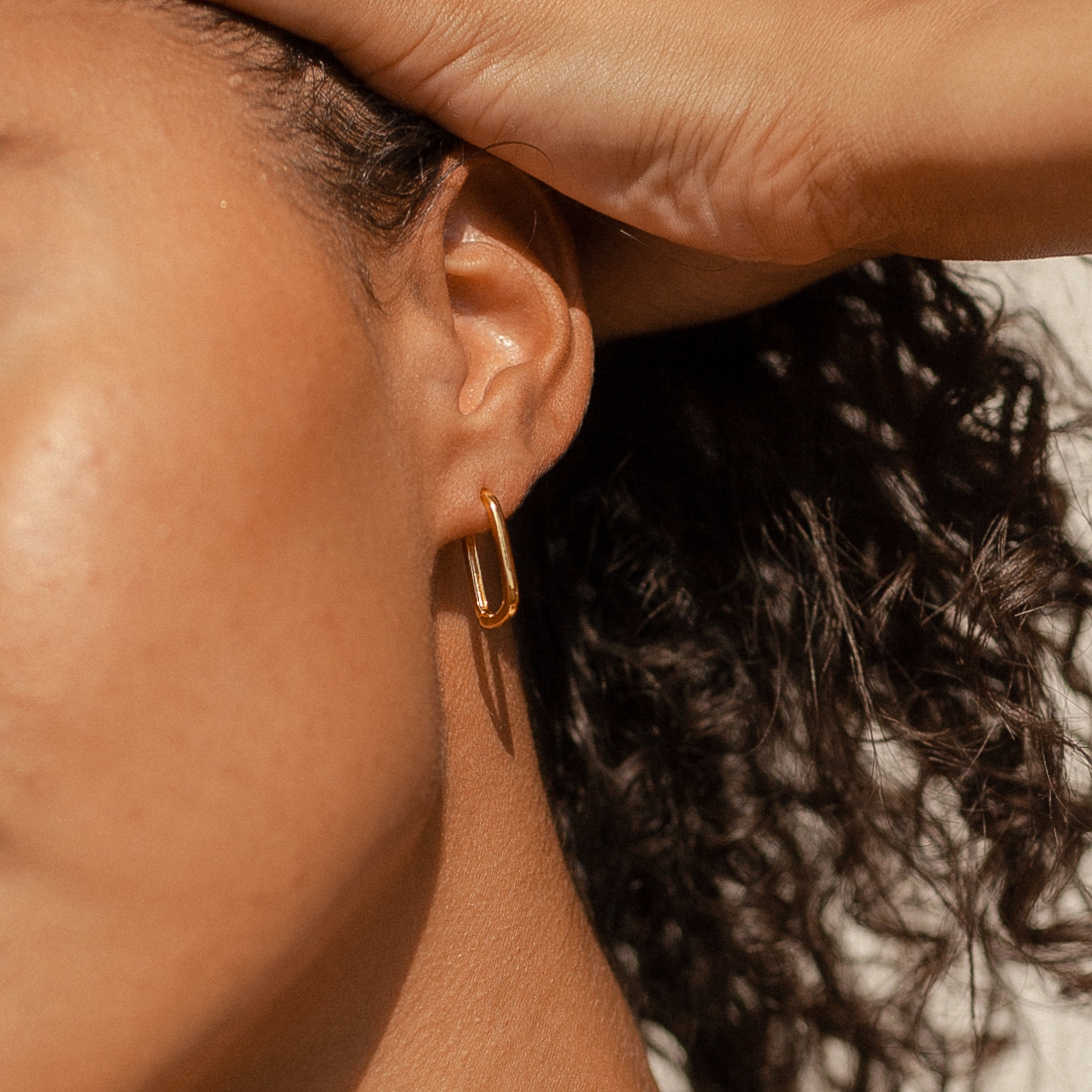 Close-up of a person’s ear wearing Nina Link Earrings in Rose Gold, with a hand resting on their head and curly hair visible.