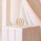Classic Hoops are chic gold midi hoop earrings, featured on a white textured pedestal with a soft beige fabric background—an elegant choice for the modern city girl.